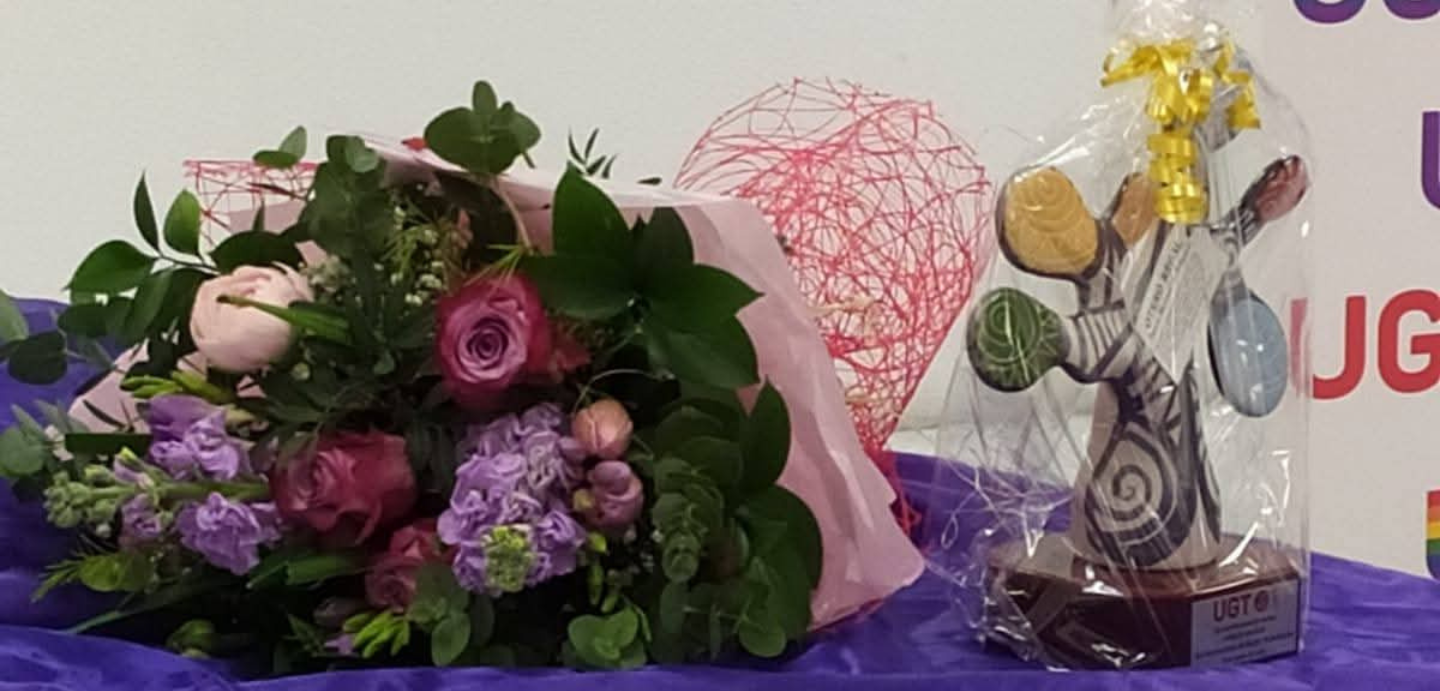 Imaxe dunha mesa onde está colocado o premio "Distinción muller traballadora" e un ramo de flores.