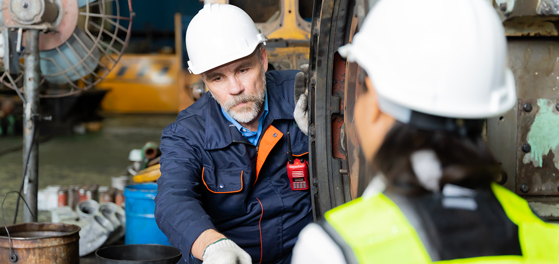 Dúas persoas técnicas que levan posto un casco de cor blanca e un equipo de seguridade traballando nun espazo industrial.